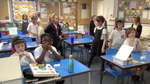 Children are wearing pink headphones.  Some are sitting at tables and some are walking around the classroom.