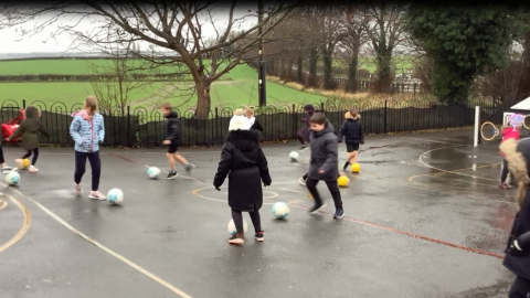 A group of children are practising close control of a football on a rainy playground.