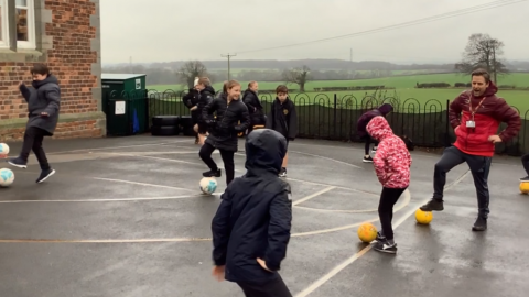 Children dressed in PE kit and coats on a rainy playground.  They have one foot on a football and their hands on their hips.  They are smiling.