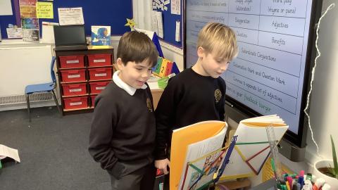 Two boys are reading from their books propped up on a music stand.  They are in front of an interactive whiteboard.