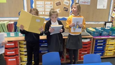 Children are reading from their books near to some drawers in a classroom.