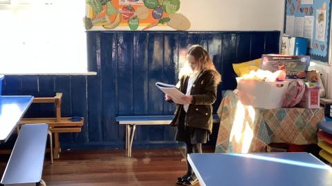 A child stands in the corner of the school hall reading from a book.