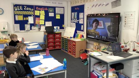 Children are sitting at desks looking at another child's image on the whiteboard form a video link.