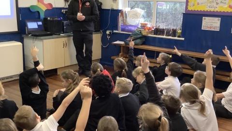 Fire Safety office talking to children in a school hall. CHildren have their hands up.