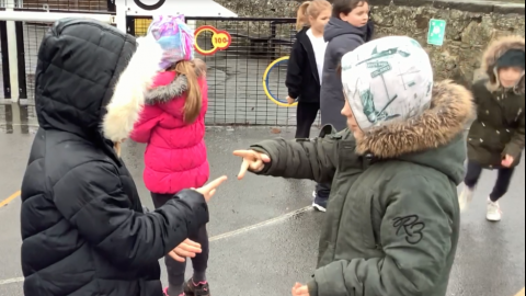 A close up of 2 children playing rock, paper scissors as part of their PE warm up.  In the background a child is running while others look on.