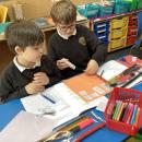 Two biys work together on coloured paper sticking a hundred square which has been cut up into sections