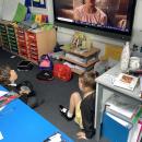 A boy and a girl sit on the floor in front of an interactive whiteboard.  There is an image of girl on the screen.